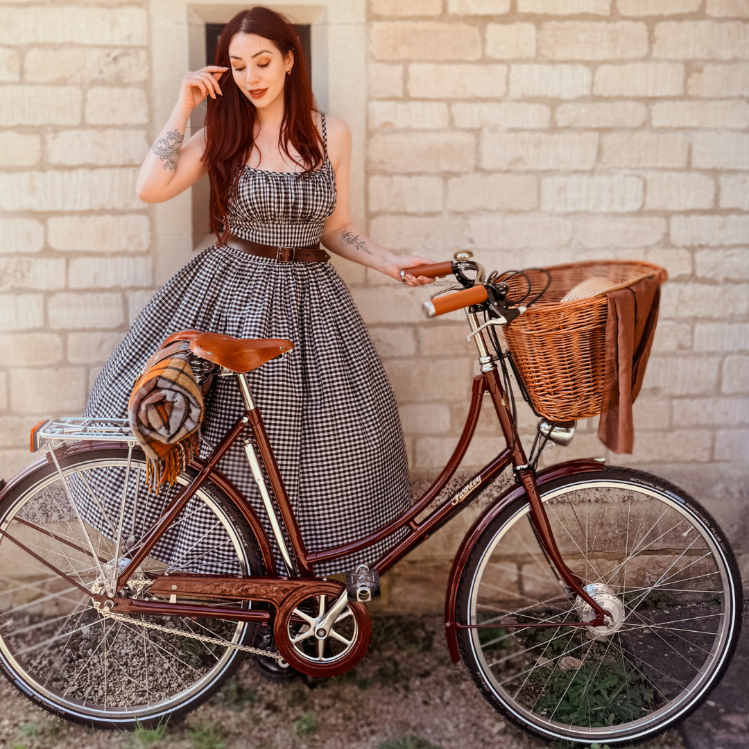 A lady with long red hair wearing a gingham dress stood behind her burgundy classic Britannia bicycle with front wicker basket