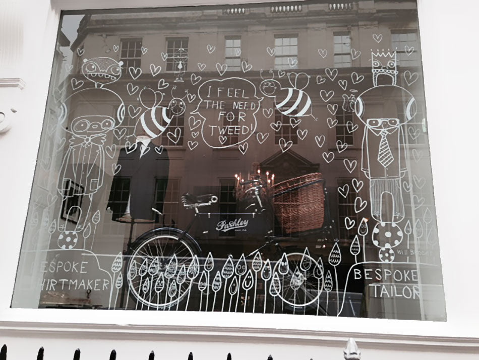 A window display at Norton & Sons on Saville Row, London. It features a Pashley deli bike, with a large front wicker basket, and with jolly illustrations drawn in white on the window's glass.