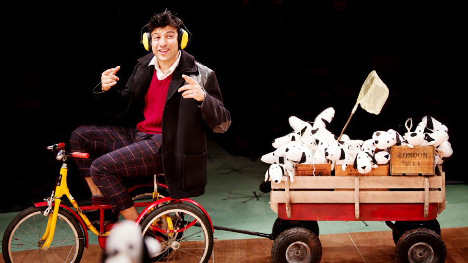 A man with ear defenders riding a Pashley Pickle tricycle pulling a cart full of 101 Dalmatian soft toys.