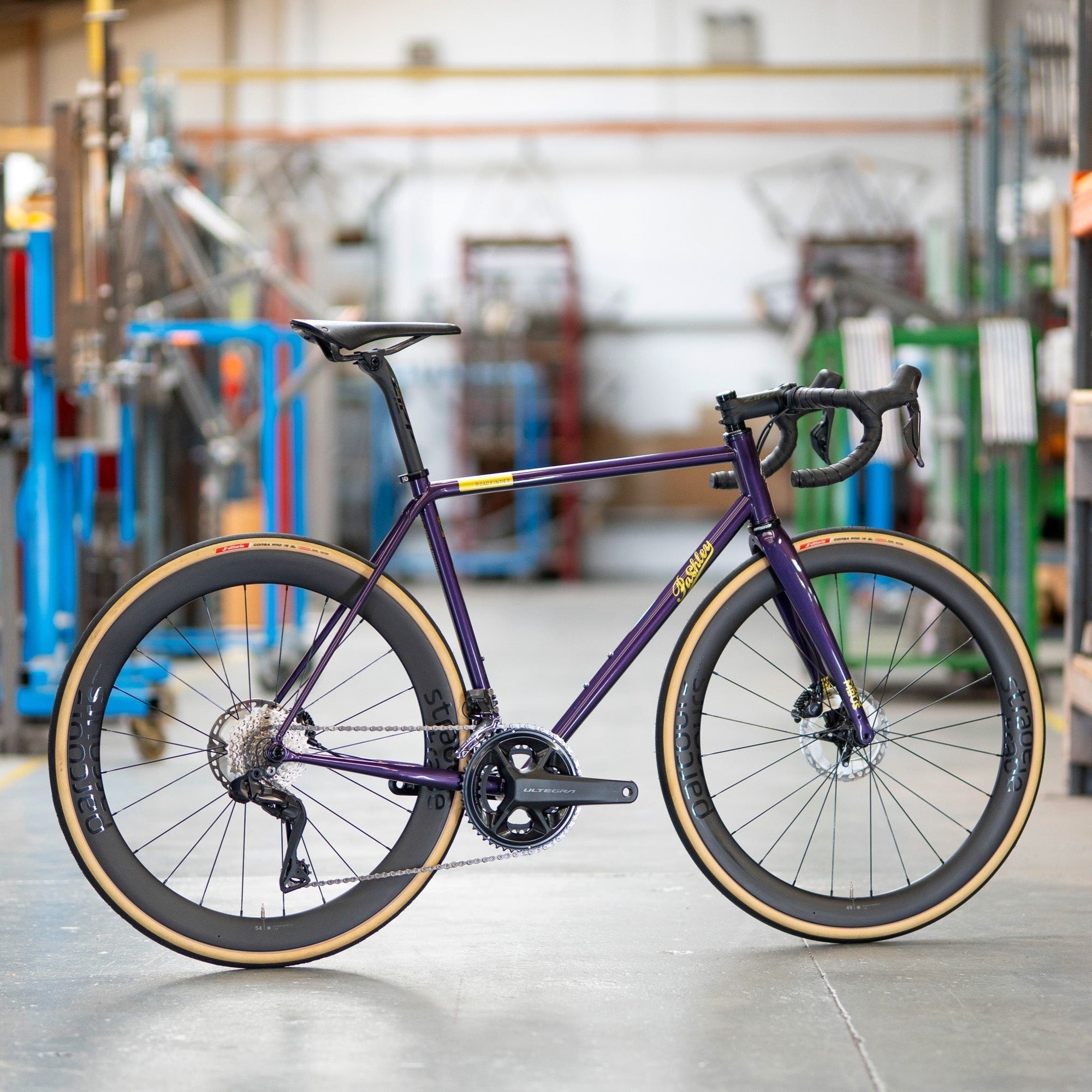 A purple Pashley Roadfinder SL road bike pictured on the grey factory floor with steel bike frames behind