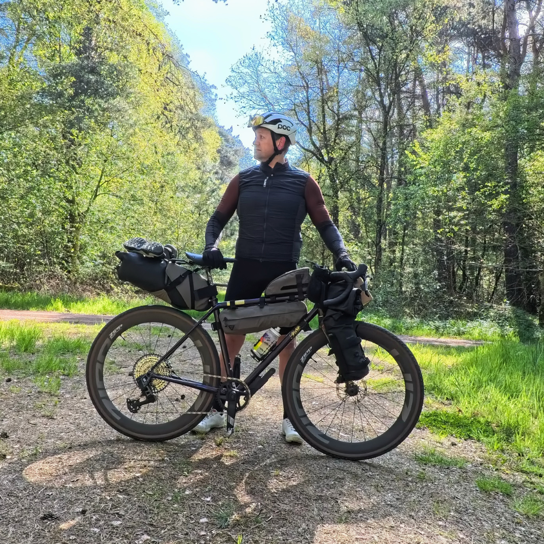 A ride in a black jersey and white helmet stood behind his Roadfinder X gravel bike on a gravel track. The bike is loaded with bags and kit and there are trees and blue skies behind