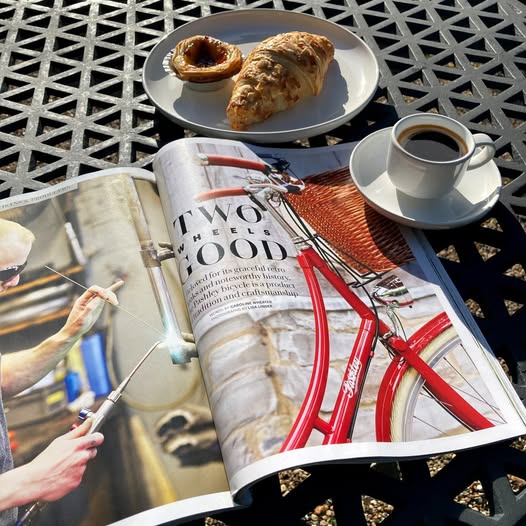 An open magazine featuring a red Pashley classic bicycle with front wicker basket, on a table with a white coffee cup and a plate of pastries