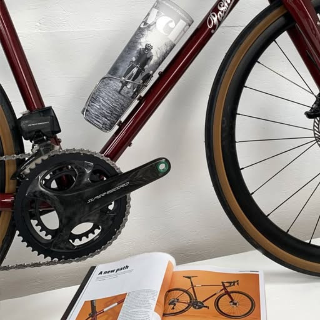 An magazine rolled into the bottle cage on a burgundy Roadfinder bicycle, with an open magazine in front