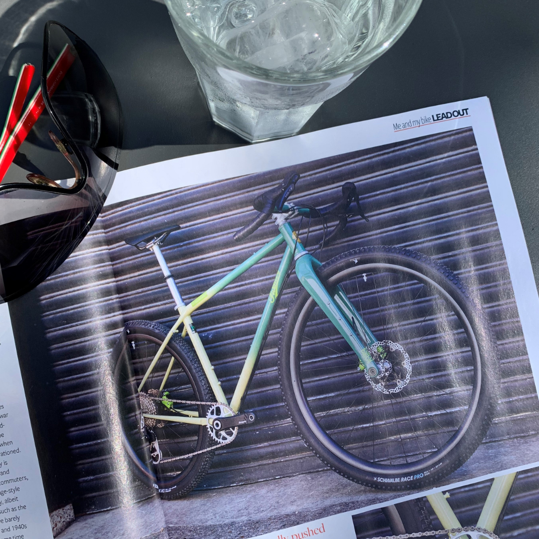 Open magazine on a table beside a glass of water and a pair of sunglasses. The magazine shows a photo of Pashley's Wildfinder Mullet built for Bespoked.