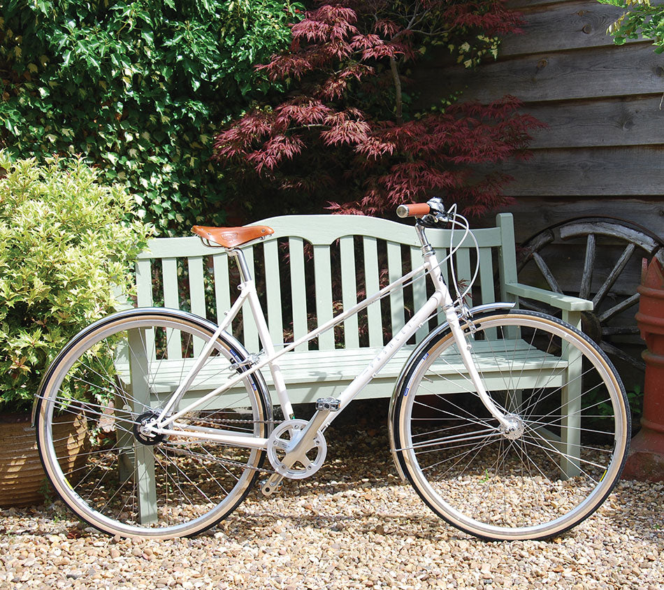 An old English white mixte Aurora bicycle standing next to a duck-egg bench and on top of gravel in a leafy garden.