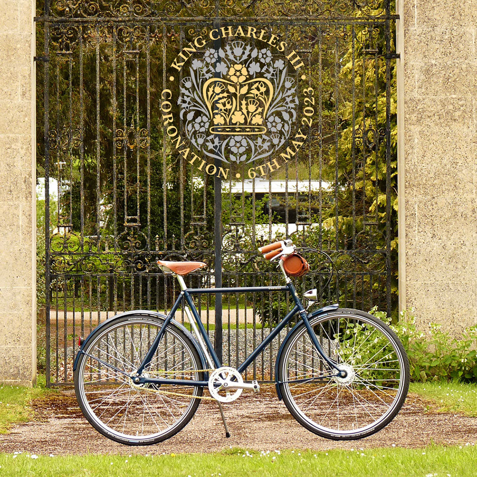 A blue classic gents bicycle with leather saddle and bar bag, parked in front of stately iron gates.