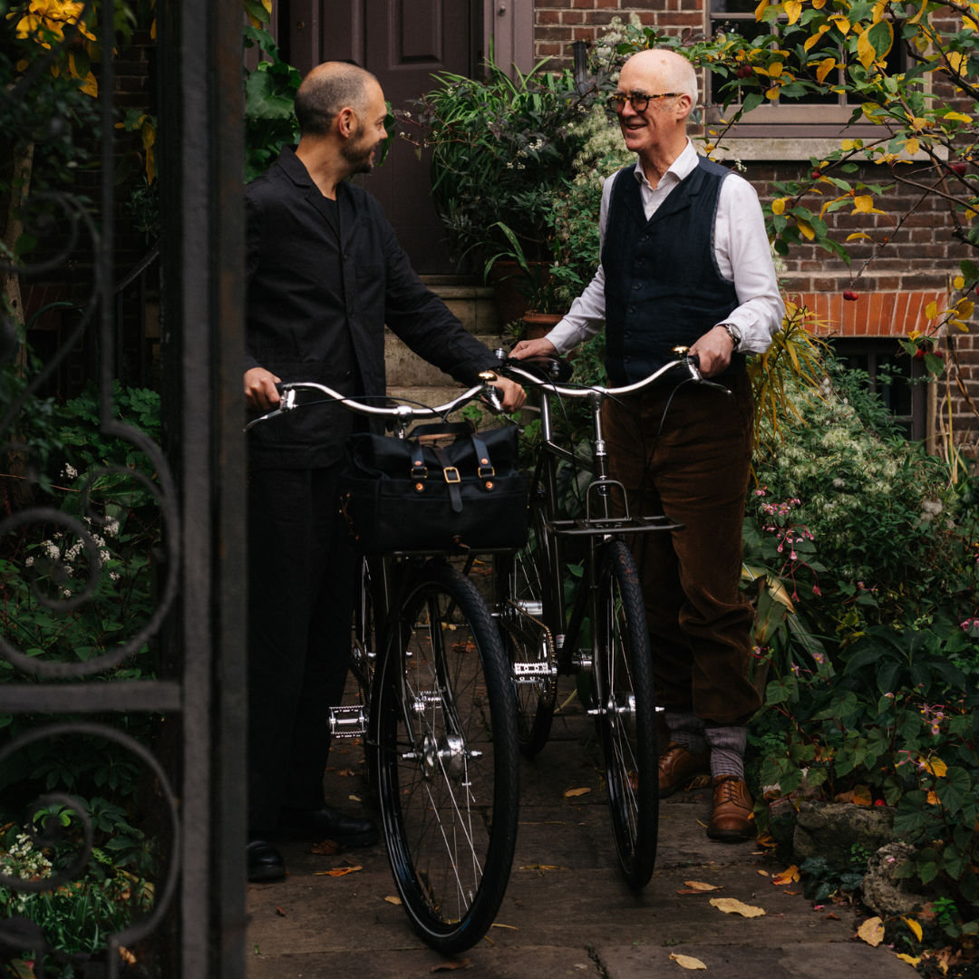 Two smartly dressed gents stood holding their classic bicycles in a garden with a townhouse behind