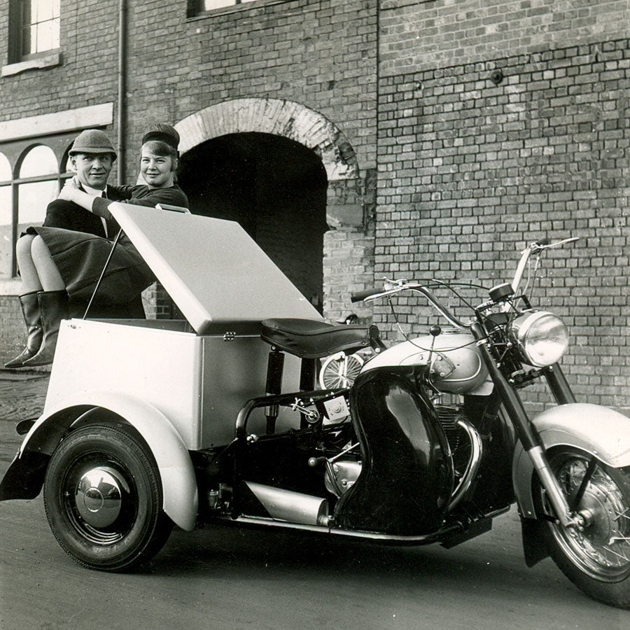 Man pretending to lift a lady into the cargo box of a vintage 1950s motorised tricycle, outside the old Pashley factory.