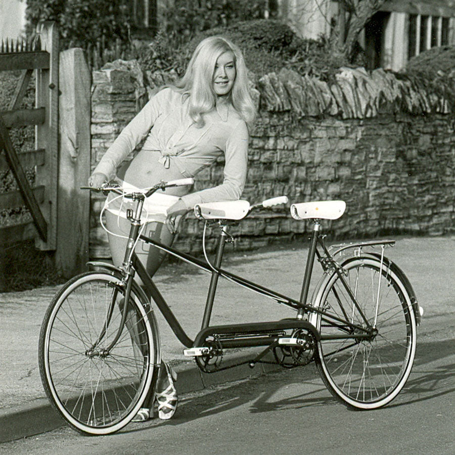 A blond lady wearing shorts and leaning on the saddle of a Pashley Tourmaster tricycle in front of a stone wall.