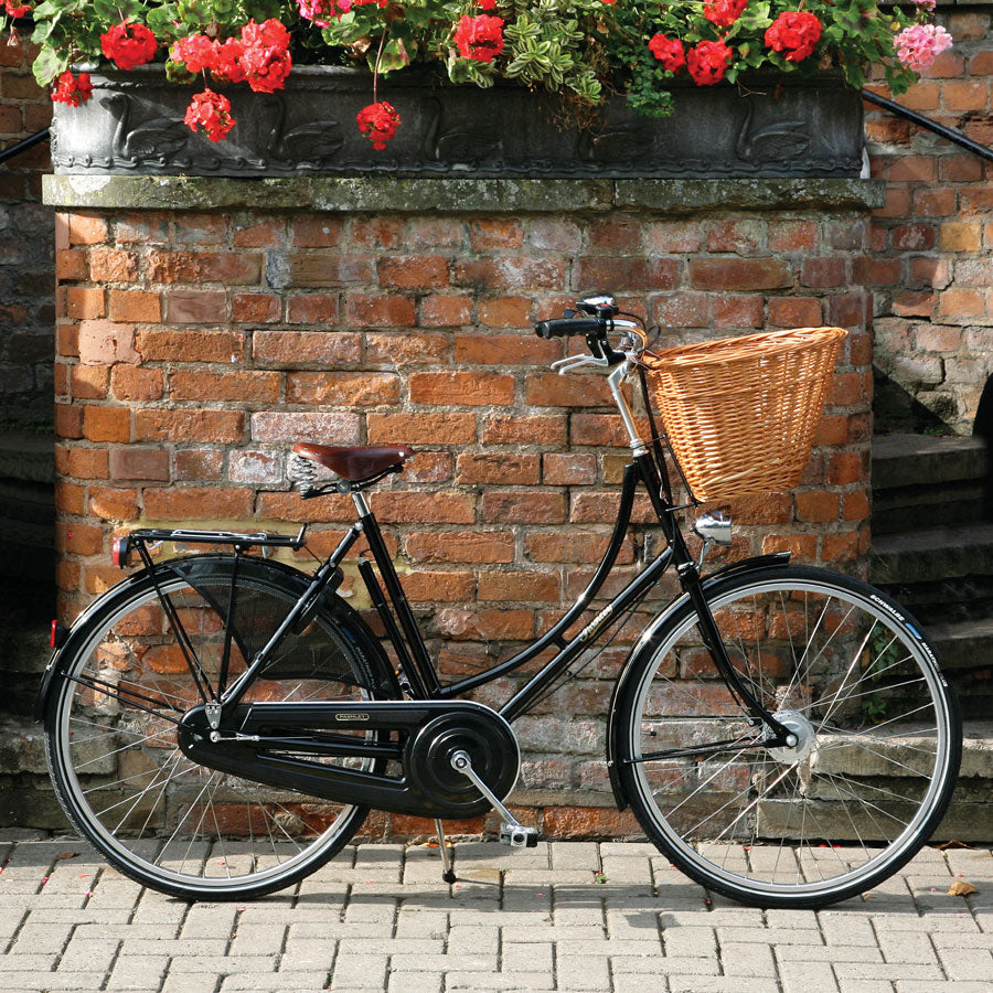 A black classic Pashley bike with wicker basket sitting next to a brick wall with red pelargoniums handing above in a flower box.