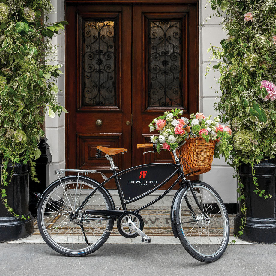 A Parabike fleet in black with a front basket full of flowers, and a frame nameboard displaying Brown's Hotel's logo. The bike is propped outside the hotel's front entrance.