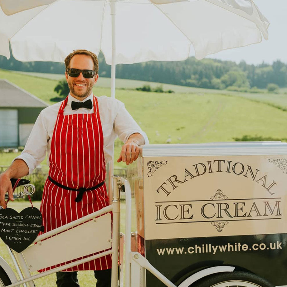 An ice cream seller in a red stripped apron, next to a cream coloured classic ice cream vending trike with branding on its freezer.