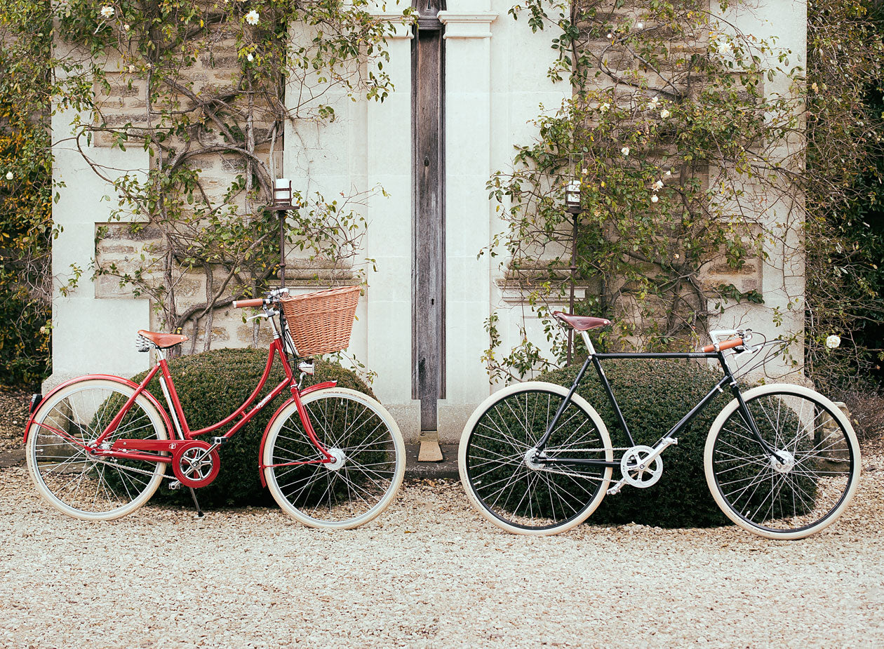 A womens red classic bicycle with basket next to a mens vintage racing bike in front of an old house with climbing roses.