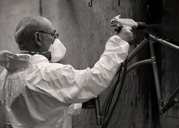 A man wearing a mask and overalls spraying a powder coat paint on to a classic bicycle frame.