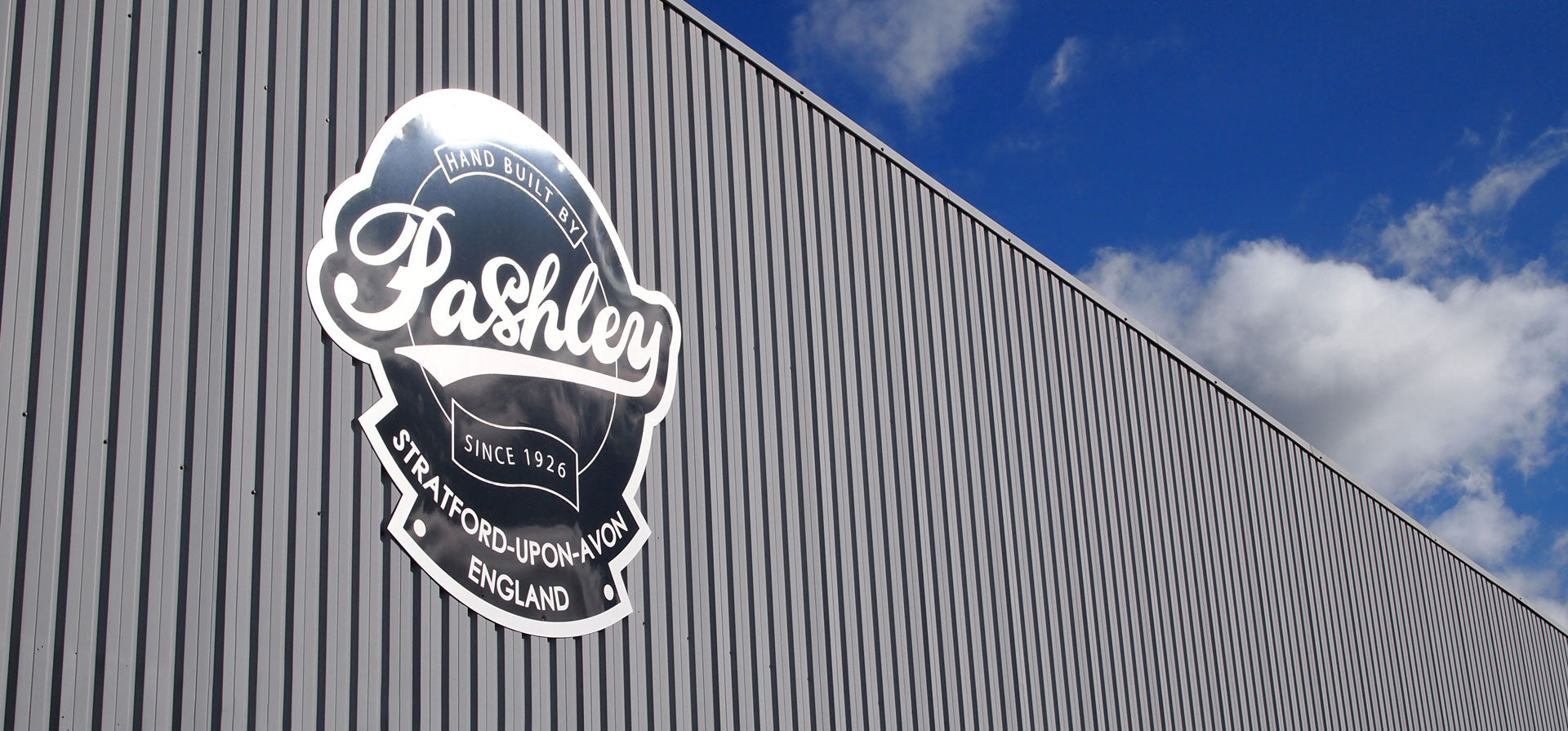A large Pashley headbadge sign on the front of the Pashley bike factory with blue sky and clouds in the background.