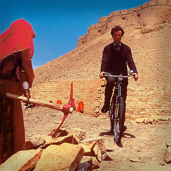 Gentleman riding a black roadster bicycle across sandy desert next to women with red head scarf breaking rock with an pick axe