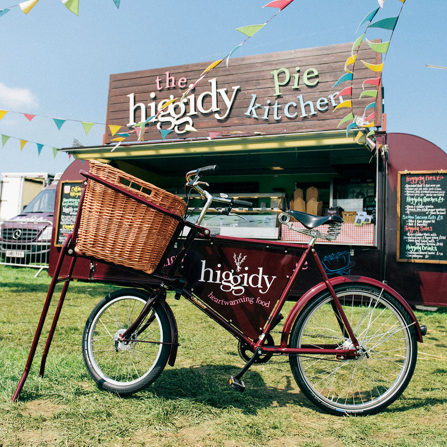 The Pashley butcher's style deli bike in a dark red colour with higgidy pies branded frame plate, sitting in front of their mobile kitchen.