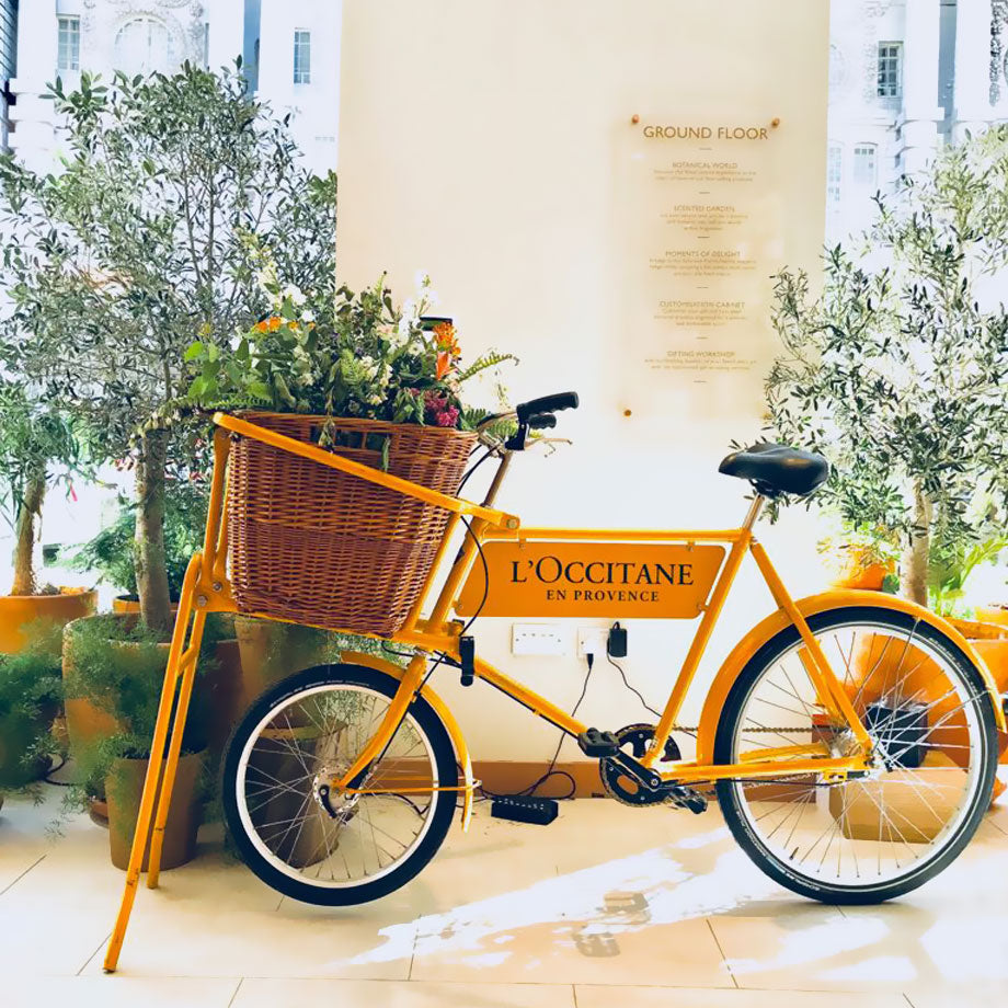 Yellow deli bike with larger wicker basket full of wild flowers and with l'occitane logo on the bicycles advertising board.