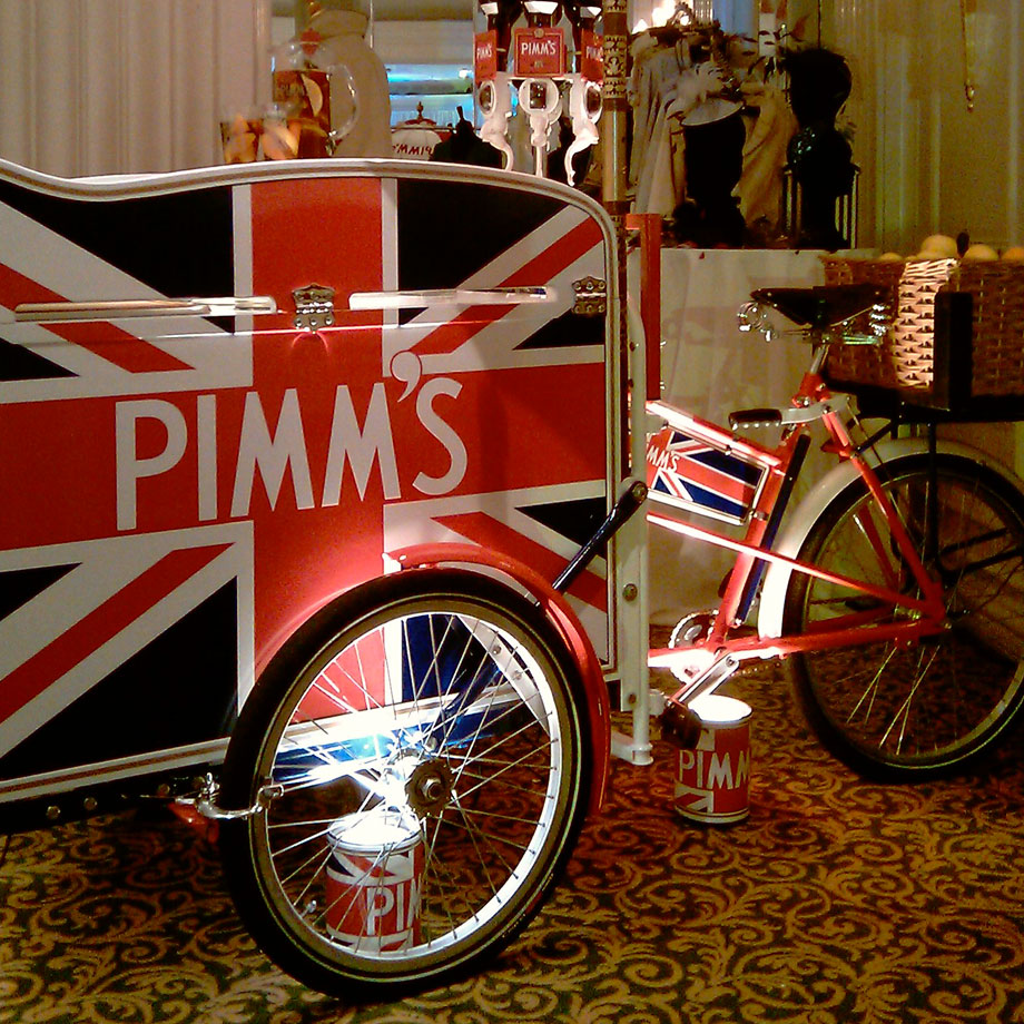 A Pashley classic vending trike with large front cool box, printed with PIMM's branding, with mini bottles for serving attached around it's umbrella pole.