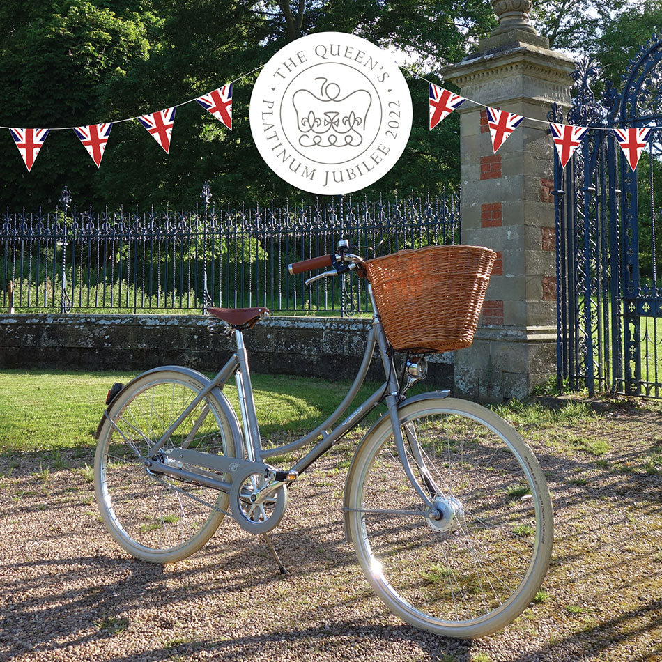 A silver Britannia bicycle with a large front wicker basket, sitting beneath union jack bunting and in front of Charlecote Park front gates.
