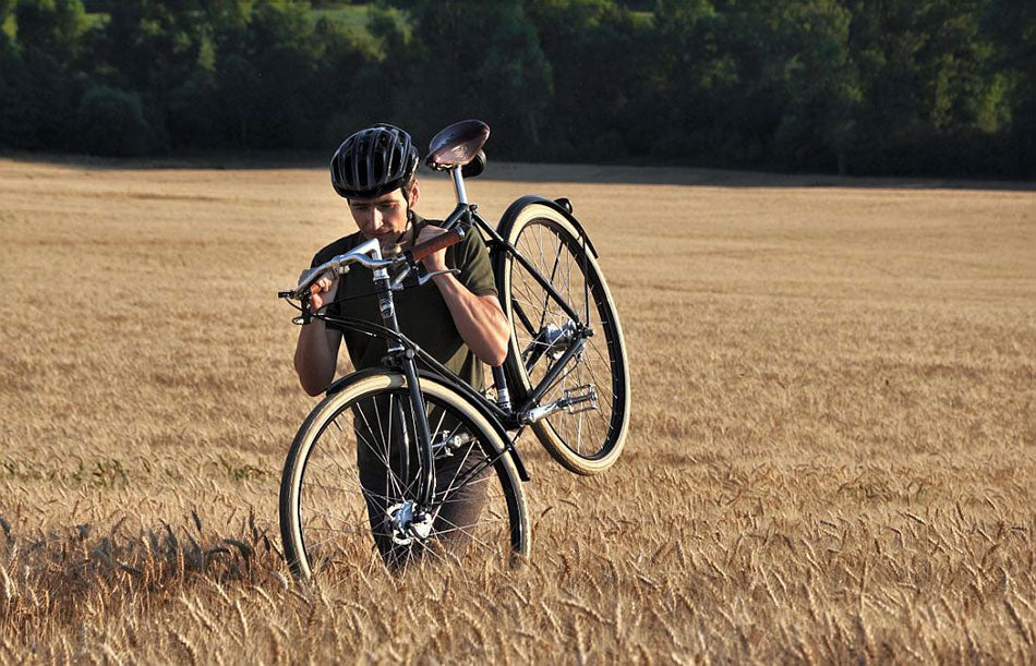 A man walking through a wheat grain field in Slovakia carrying a Pashley Guv'nor bicycle over his shoulder.