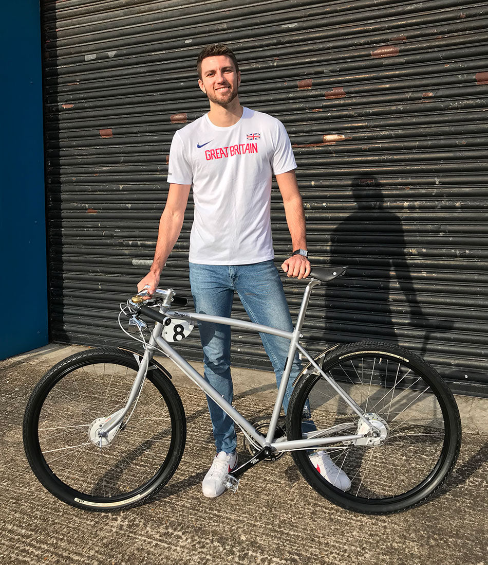 Olympic hurdler, Andrew Pozzi, wearing a Great Britain top, stand with a Pashley-Morgan 8 speed in front of a black roller door.