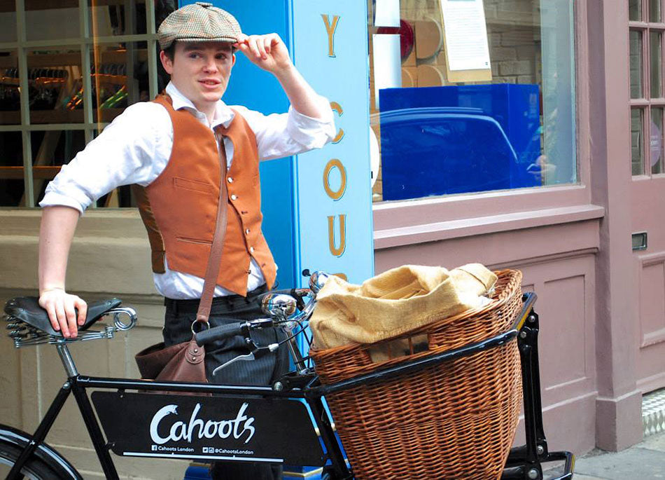 A young gent in a flat cap and tailored waistcoat, tipping his hat whilst standing next to his Hovis style Pashley deli bike.