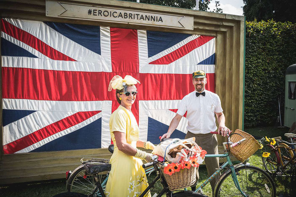 A lady in a yellow vintage dress standing with her classic bike and alongside her partner with his bike. Their is a very large union jack flag behind them.