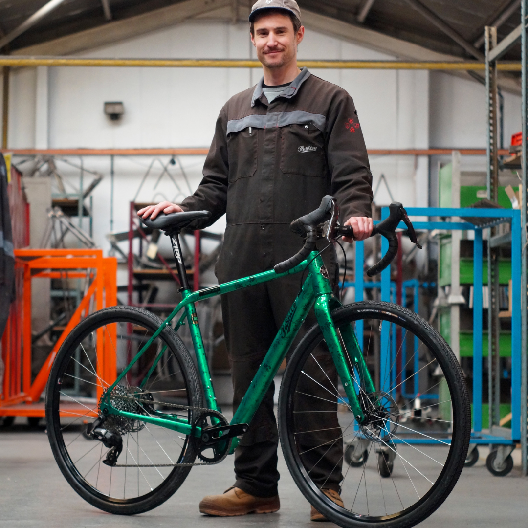 A craftsman in black overalls stood holding an emerald green metallic Roadfinder XE bike within the Pashley factory