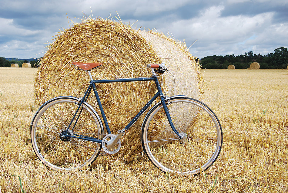 A dusk blue classic steel frame Pashley countryman bike leaning against a large, rolled hale bale in the Cotswolds.