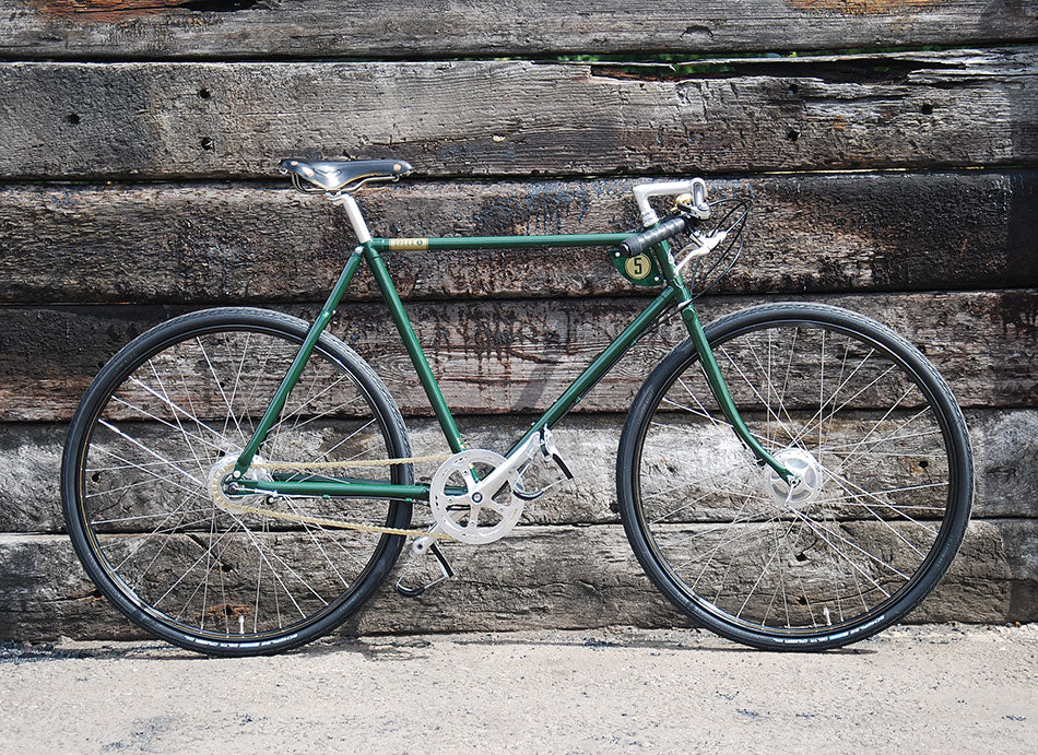 A British racing green classic path racer bicycle with black leather saddle and gold bicycle chain. It is leaning against a wall made from old, grey railway sleepers.