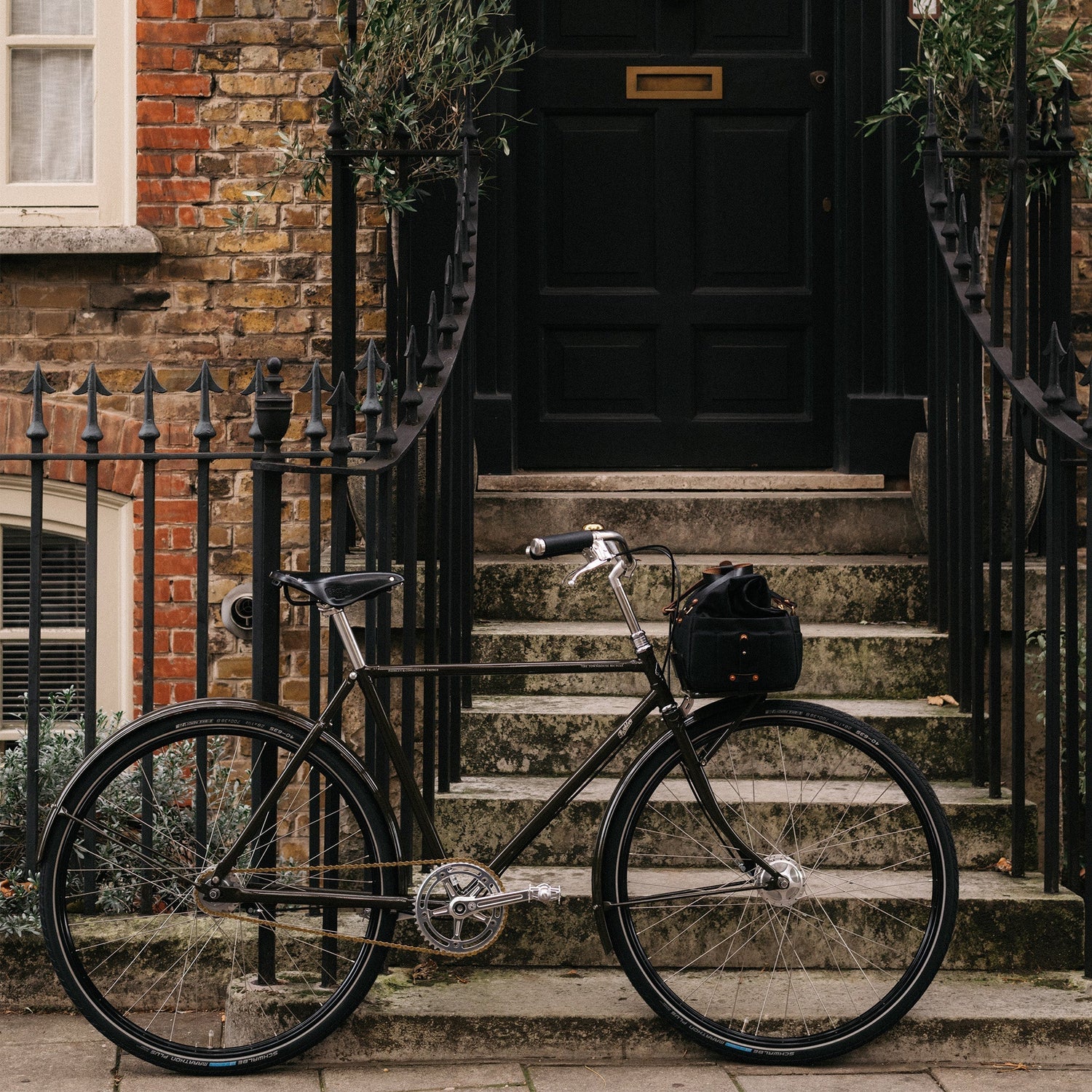 Black classic TownHouse bicycle with a black bag on the front carrier, against black railings in front of a townhouse, with steps leading to a black door