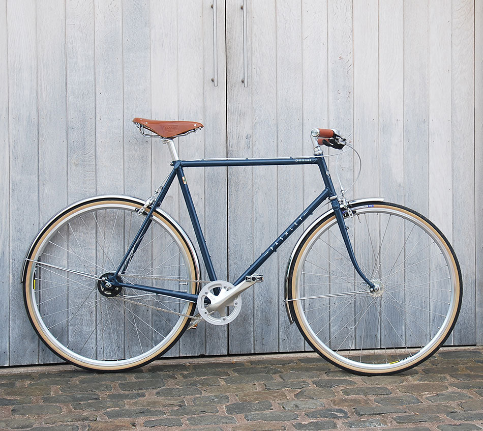 A dark dusk blue, lugged steel Countryman bicycle with brooks leather saddle and tan wall bike tyres. It is leaning against ash grey barn doors.