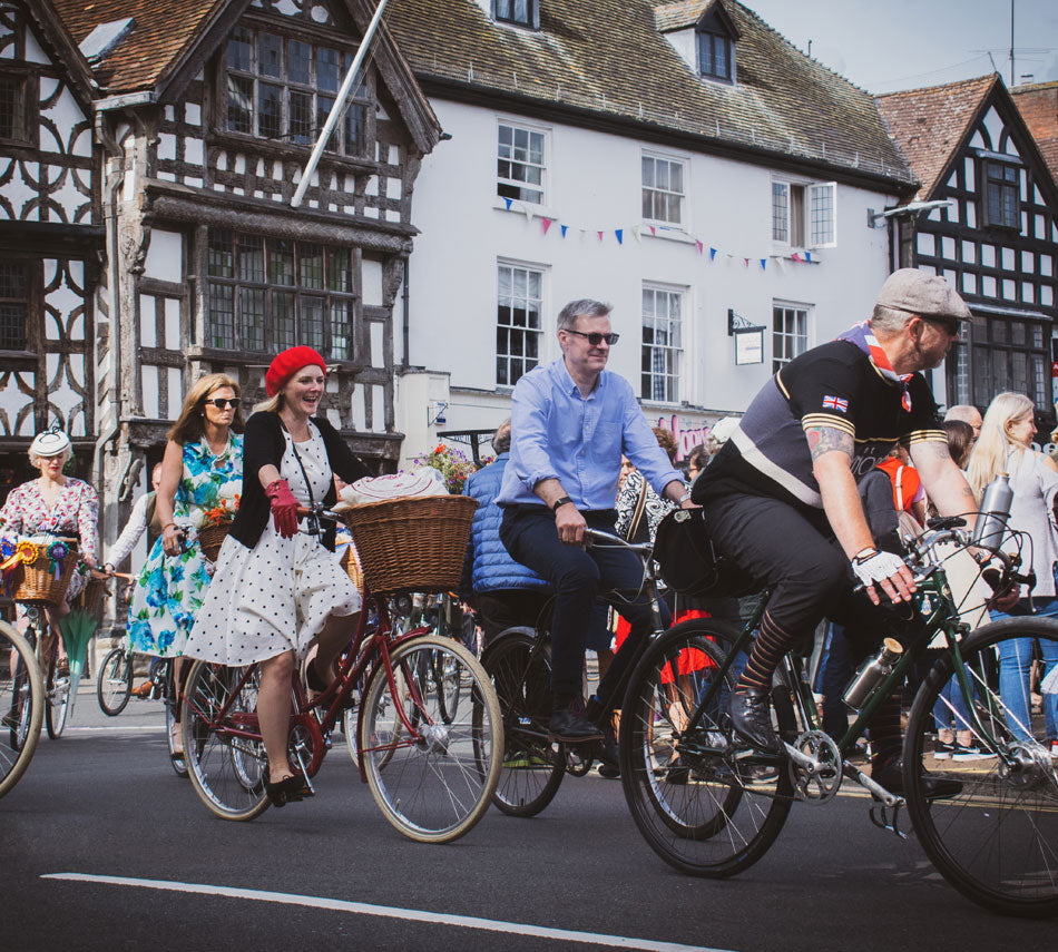 A peloton of Pashley cyclists riding through the streets of Stratford-upon-Avon, dressed in pretty vintage dresses or retro cycle clothing.