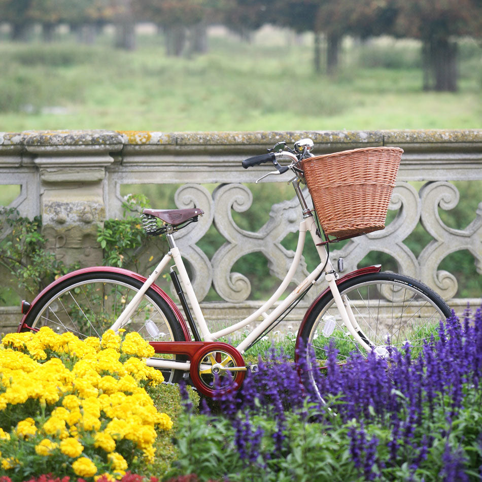 Pashley ladies Sonnet bicycle in crema and red next to an old stone wall and a flower bed of yellow and purple flowers