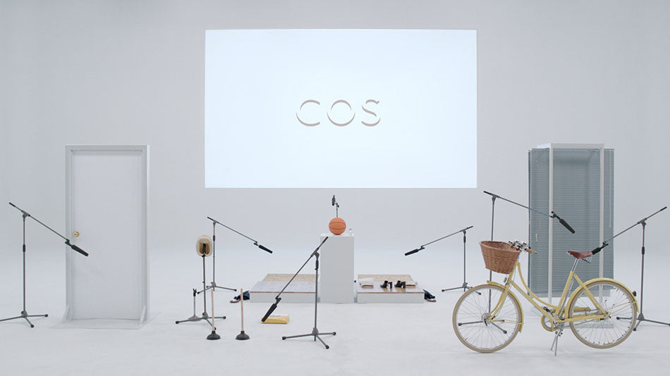 A white studio with projection of the COS brand logo on the wall with mic stands, freestanding doors and a yellow Pashley Poppy standing in front.