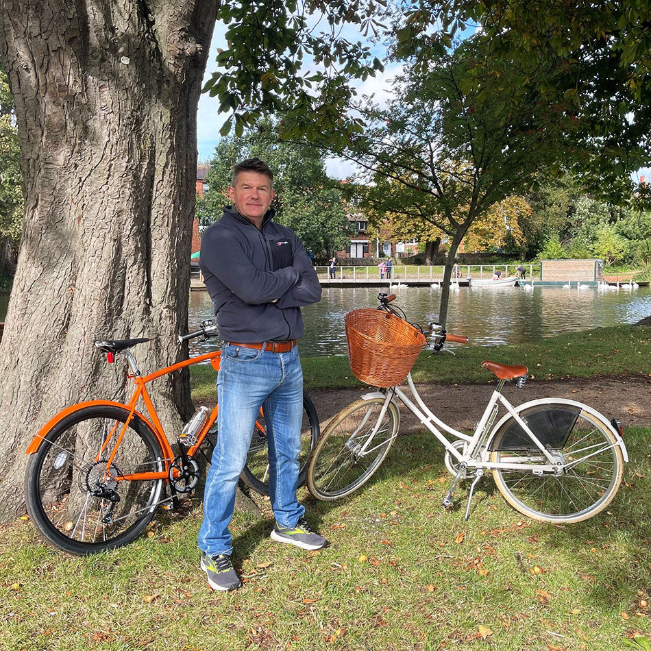 Man standing by the river Avon and next to a bright orange classic racer bicycle and a white vintage bicycle.