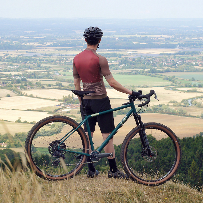 A rider in a red jersey and black helmet with his back to the camera taking in a view of a valley from a hilltop, stood next to his Tempest Blue Wildfinder gravel bike