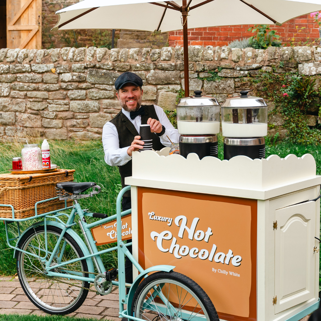A gent in a waistcoat and flat cap stood behind his Pashley vending trike making luxury hot chocolate