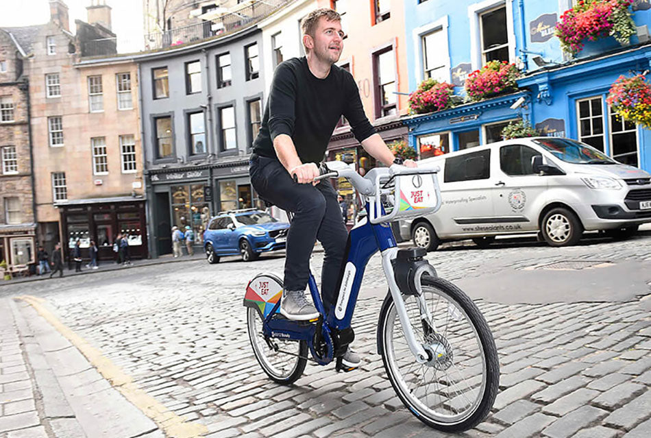 A man in a black t-shirt cycling up Victoria Street on a blue Just Eat branded Edinburgh city hire e-bike.
