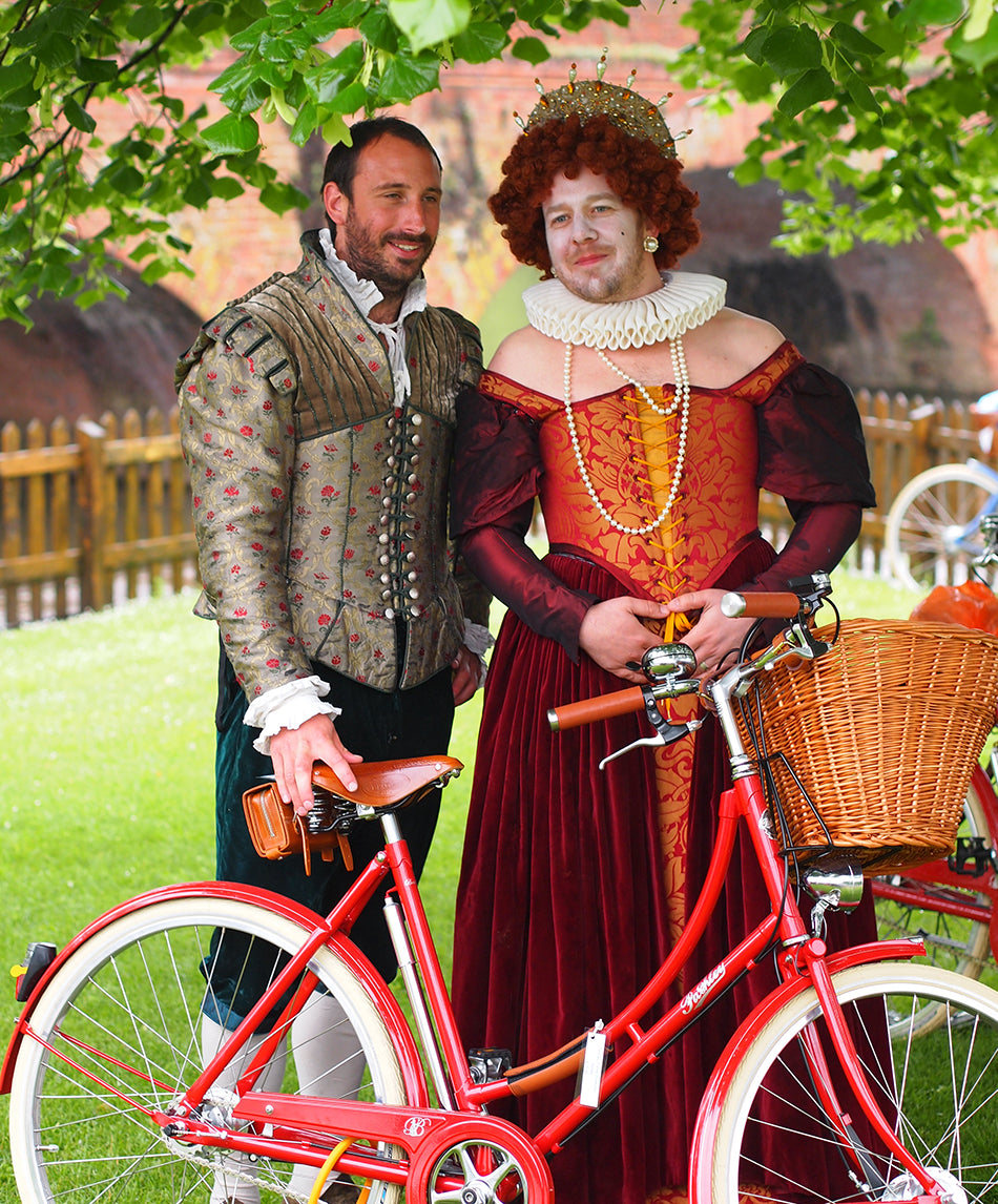 A gentlemen dressed as a Shakespearean Queen alongside a companion and a Britannia Bicycle in red.