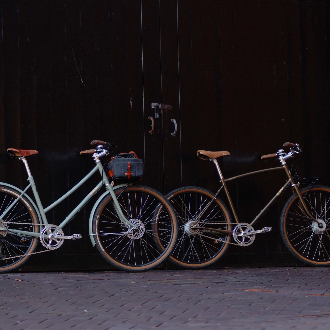 Two Prospero bicycles against a black door - one Sage with step-through frame and one Bronze with diamond frame