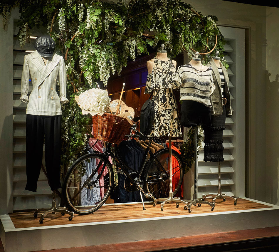A window display featuring a black Pashley vintage bicycle with wicker basket holding flowers standing alongside mannequins dressed in ralph lauren clothing.