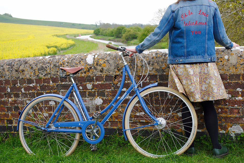 A young lady wearing an embroidered denim jacket standing next to her blue Pashley Poppy bicycle on a stone canal bridge.