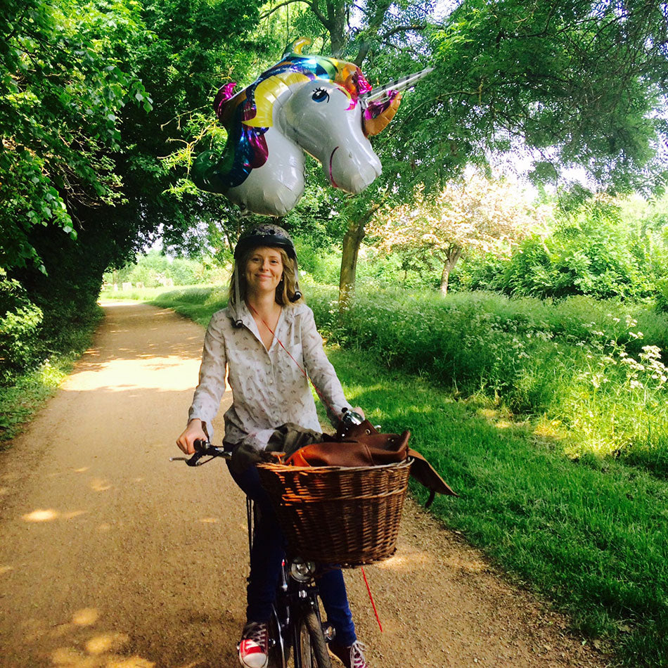 A young lady cycling her Pashley bicycle along a cycle path with a unicorn helium balloon tied behind her.