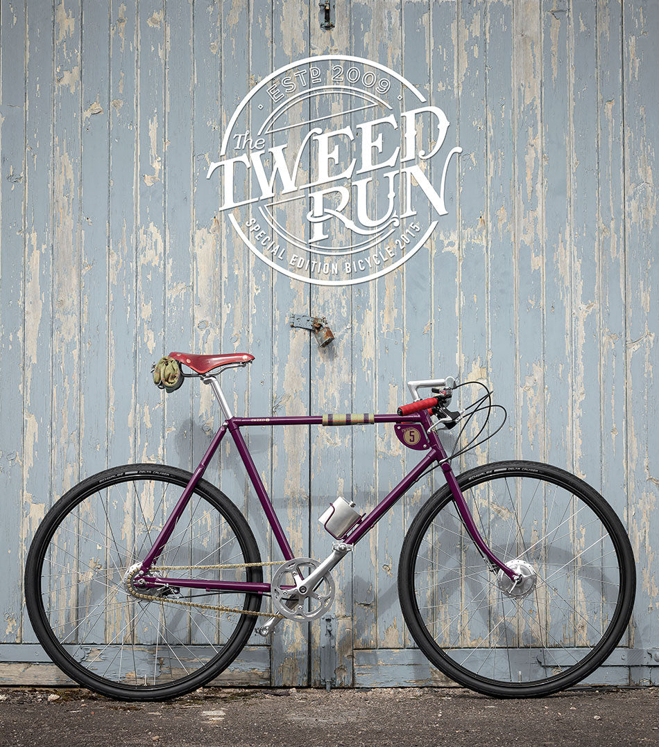 London Tweed Run Pashley 2015 bike - The TWEED 5 - in dark purple with leather saddle and reflective tweed saddle bag and frame wrap, leaning against old wooden doors.