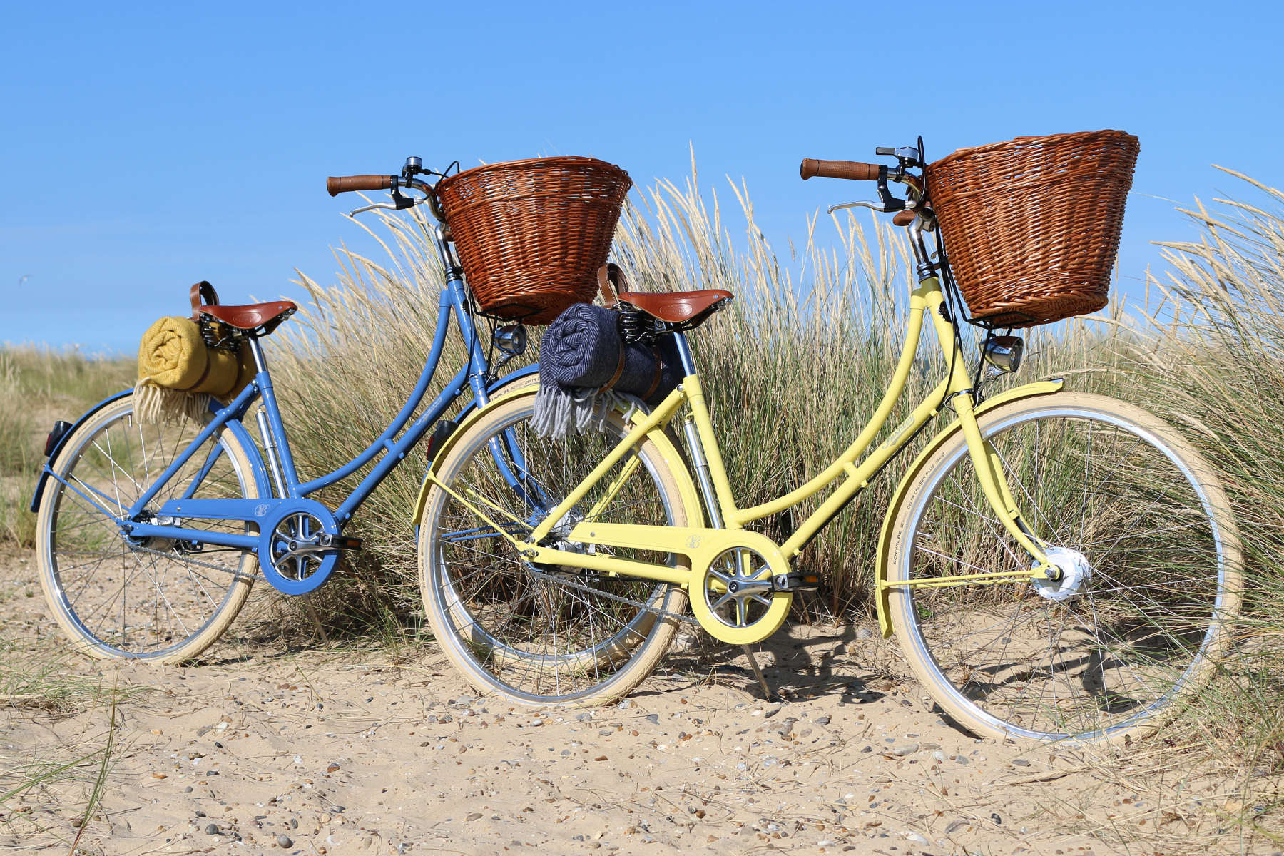 Two bicycles, one blue and one yellow, with wicker baskets on a sandy path with tall grass.