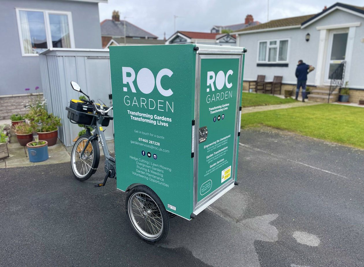 A electric cargo trike with branded rear post in front of bungalow with a gardener in the background.