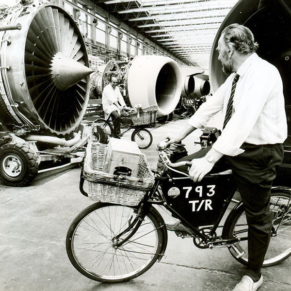 Two men sitting on 1970s cargo bicycles next to British airways turbine engines