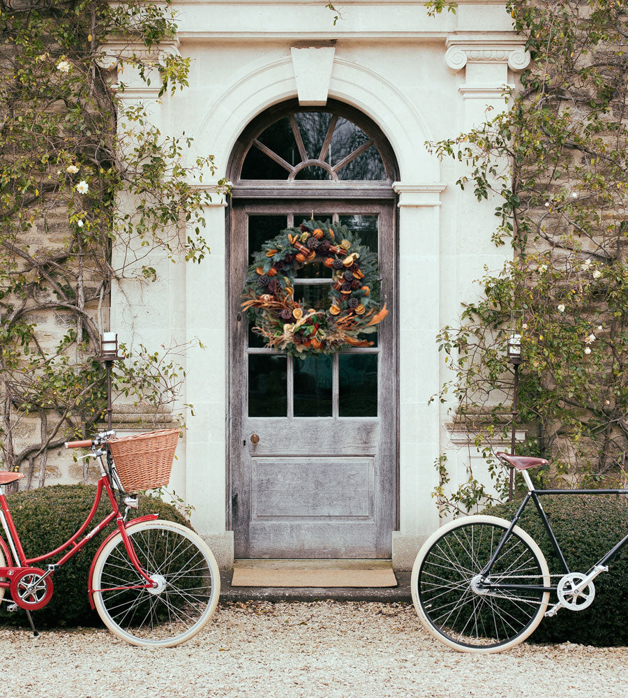 A red classic ladies bike with basket and a gentleman's black vintage racer bike sat outside a stately home front door with Christmas wreath.